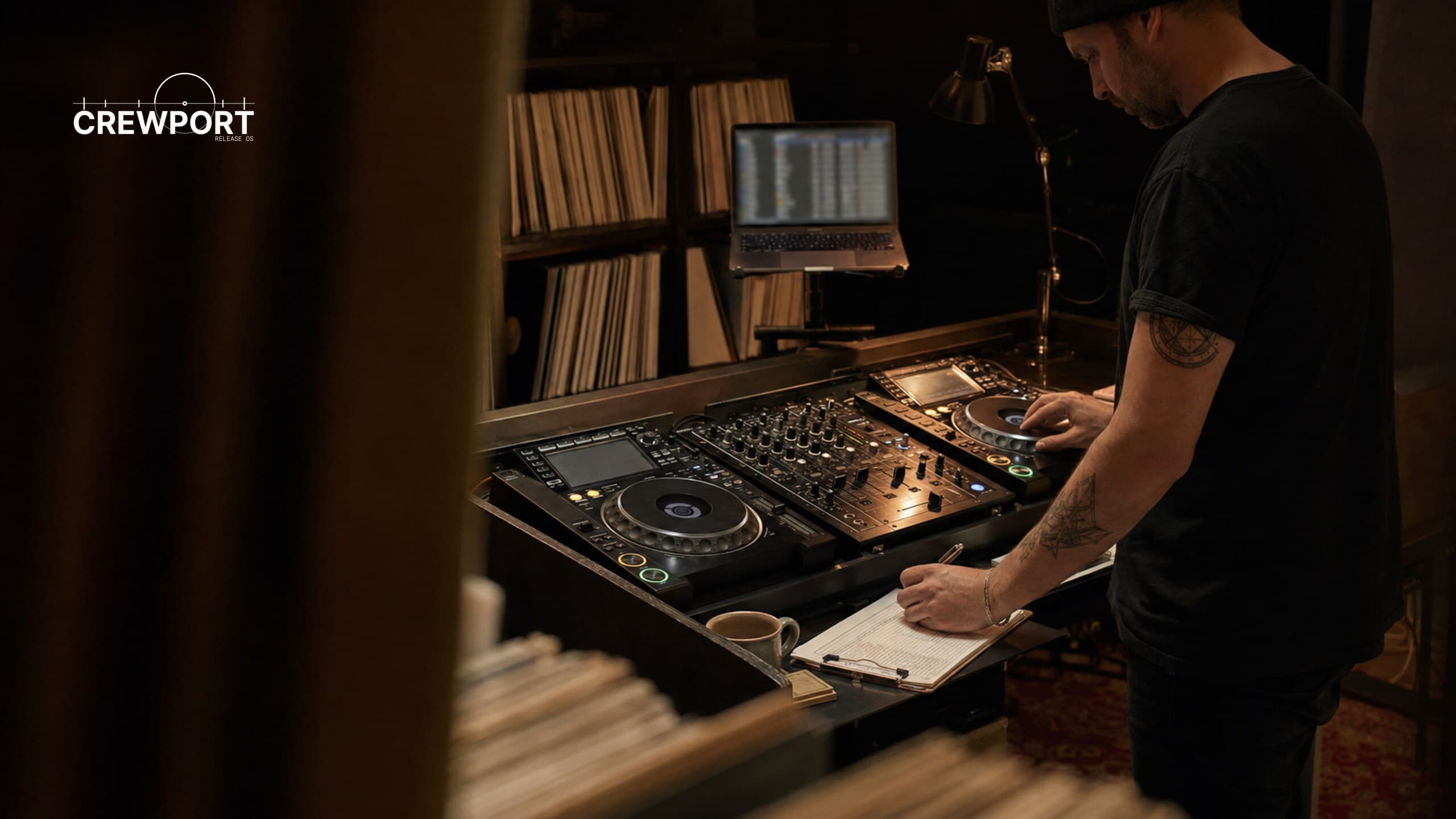 A label owner plans upcoming releases at a standing DJ setup with two decks, a mixer, laptop and vinyl collection.